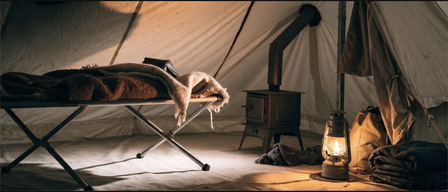 Inside a canvas wall tent with a cot, wool blankets, a small wood stove and a lantern